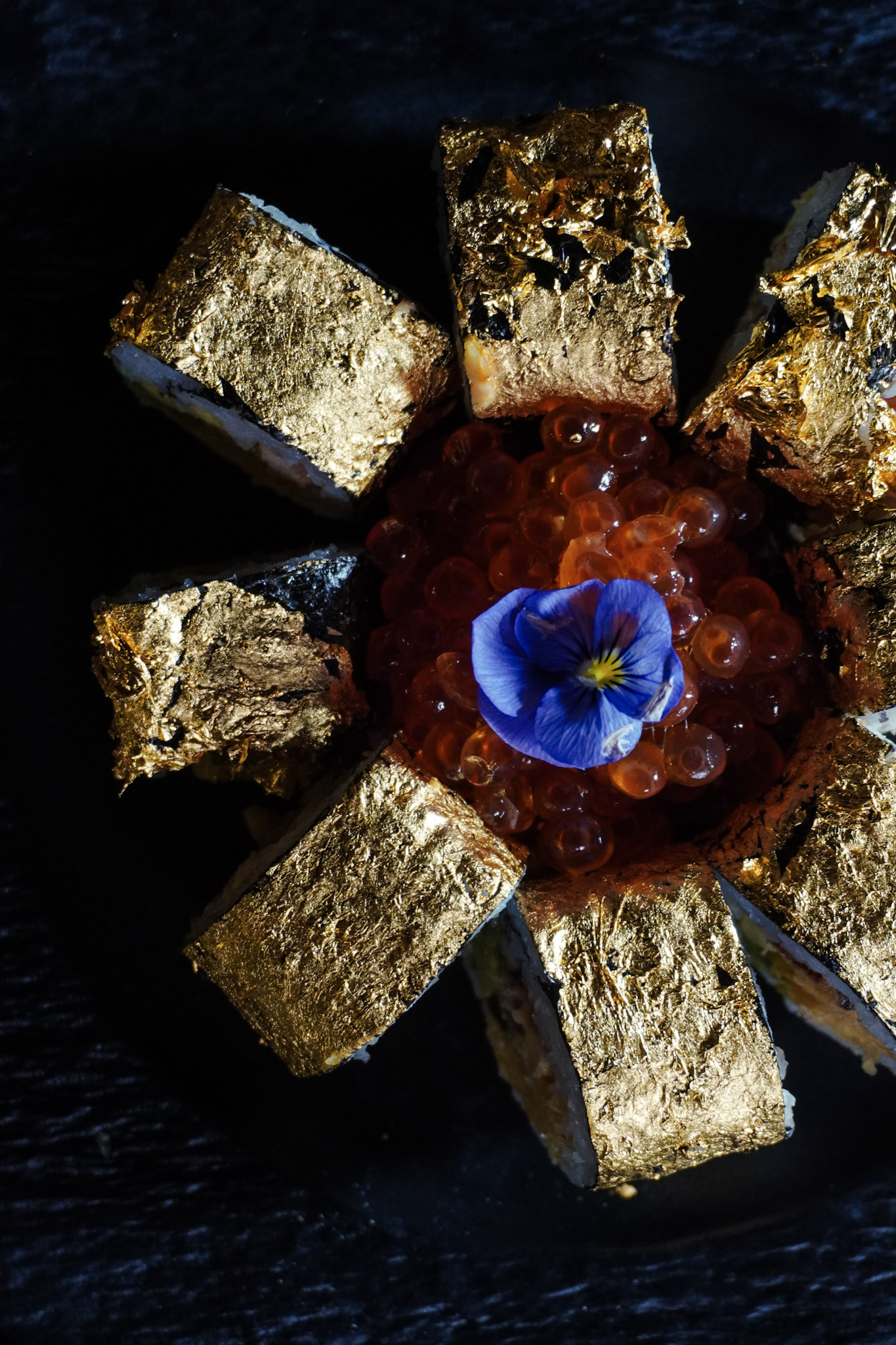 Overhead view of gold leaf sushi with ikura caviar and a violet pansy at the center.